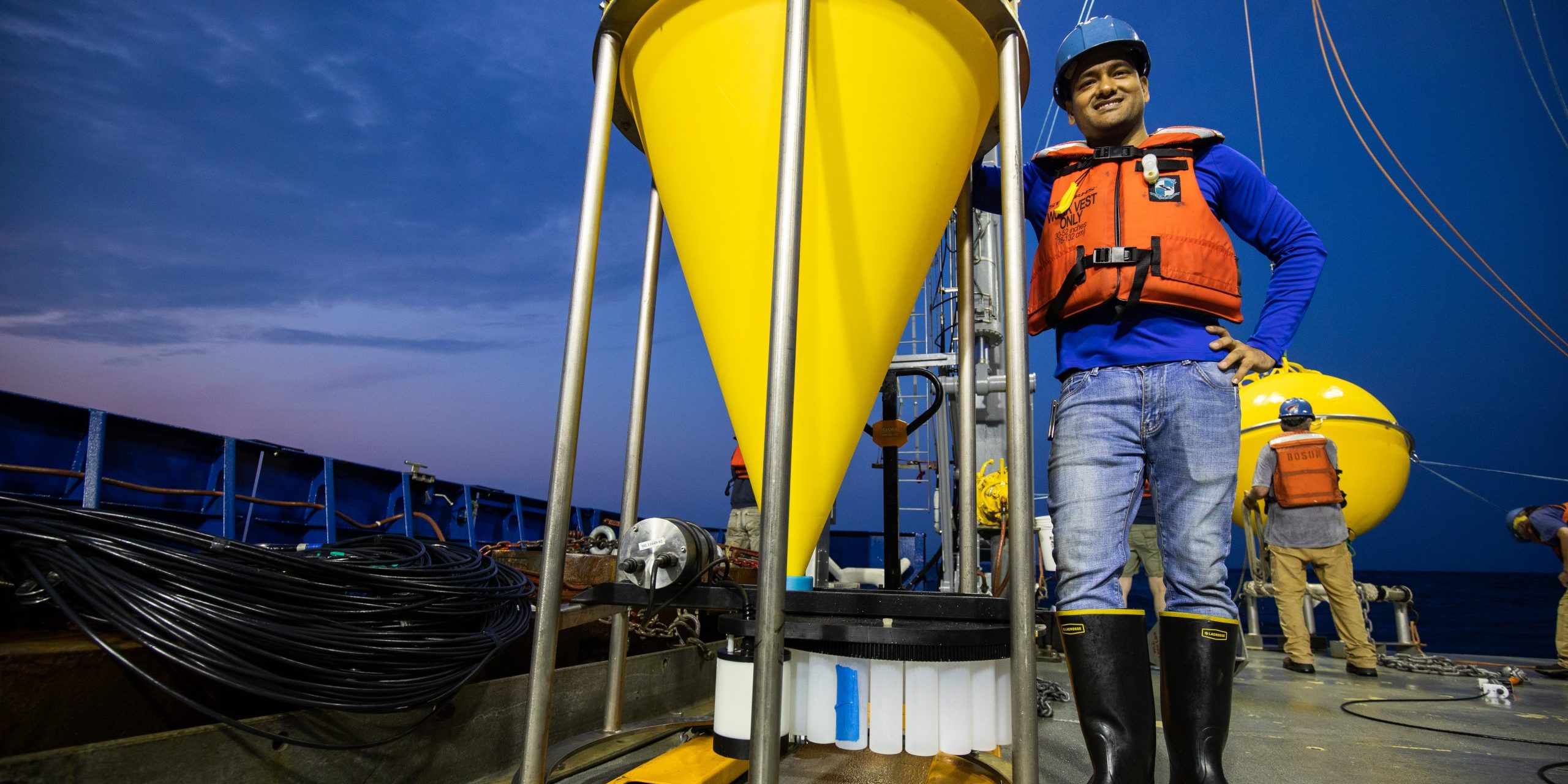 Postdoctoral scholar, Wokil Bam of Café Thorium Lab, stands tall next to a large bottom tethered trap on the stern of R/V Neil Armstrong. The trap will collect falling organic materials in the twilight zone to better understand how this layer helps the ocean sequester atmospheric carbon.